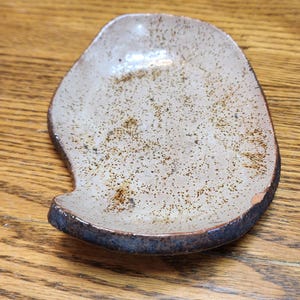 May include: A handmade ceramic dish with a unique, organic shape. The dish features a speckled, off-white interior and a dark brown rim. The exterior has a speckled pattern with brown and black spots. The dish is approximately 6 inches long.
