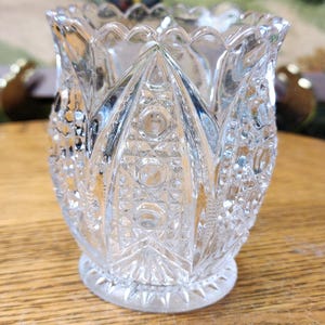May include: A clear, cut-glass votive candle holder with a scalloped rim and a detailed, geometric pattern. The glass has a textured appearance with a circular base. The candle holder is designed to hold a small candle.