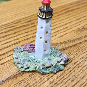 May include: A miniature lighthouse figurine with a white tower, red top, and black accents. The lighthouse has small square windows and sits on a base of green and brown textured material. The base includes a small building with a red roof.