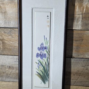 May include: A vertical rectangular wall art piece featuring a painting of purple iris flowers with green leaves on a white background. The artwork is enclosed in a dark brown wooden frame with a light-colored mat and a small metal hanger at the top.