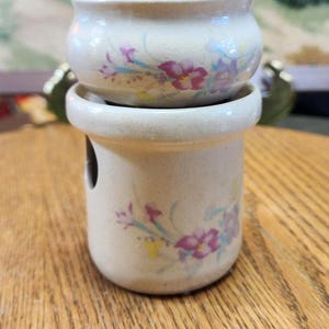 May include: A two-piece ceramic candle warmer set. The off-white pieces are decorated with purple and pink floral patterns. The warmer has a cylindrical base with a hole and a small bowl on top.