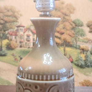 May include: A vintage brown ceramic table lamp with a decorative embossed pattern. The lamp has a bulb that is lit, and a black cord. The lamp is on a wooden table.