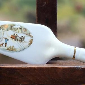 May include: White ceramic pie server with a winter scene illustration. The server features a handle with a gold accent and a hole for hanging. The illustration depicts a horse-drawn sleigh in a snowy landscape.