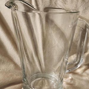 May include: A clear glass pitcher with a handle. The pitcher has a wide, angled body and a pouring spout. It is designed for serving beverages. The pitcher is made of transparent glass.