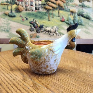 May include: A ceramic chicken-shaped container with a brown, white, and green speckled glaze. The container has a detailed tail, a white body, and a black comb. The interior is a copper color. The container is sitting on a wooden table.
