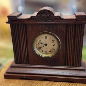 May include: A vintage wooden mantel clock with a round clock face. The clock has a dark brown finish with vertical grooves on the sides and a decorative top. The clock face has black numbers and hands.