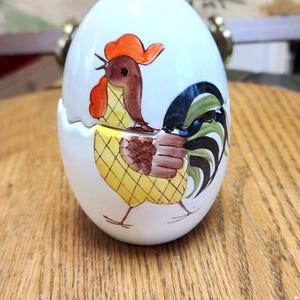 May include: A white ceramic egg-shaped container with a painted rooster design. The rooster has a red comb, brown body, and yellow and black accents. The container is on a wooden surface.