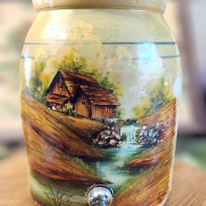 May include: A cylindrical ceramic beverage dispenser with a beige glaze and a hand-painted landscape scene featuring a brown wooden cabin, green trees, and a small waterfall. The vessel includes a silver-toned metal spigot at the base.