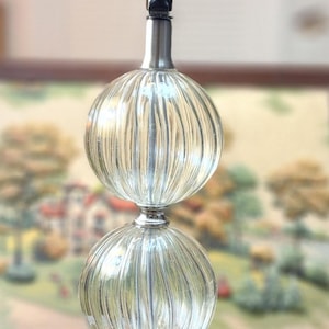 May include: A decorative table lamp with a clear, ribbed glass body and a silver metal base. The lamp features a white lightbulb and a black switch. The lamp's design is vintage and elegant.