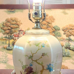 May include: A vintage table lamp with a floral design. The off-white ceramic base features a painted branch with purple and blue flowers. The lamp has a brass base and a clear glass bulb. The lamp is approximately 20 inches tall.
