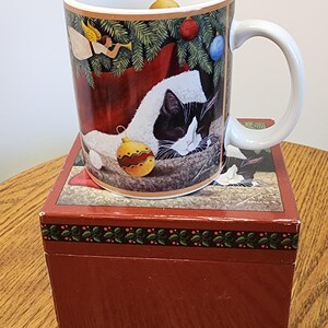 May include: A white ceramic mug featuring a holiday illustration of a black and white cat resting under a Christmas tree with ornaments and an angel. The mug sits atop a matching red rectangular box decorated with a green holly and berry border.