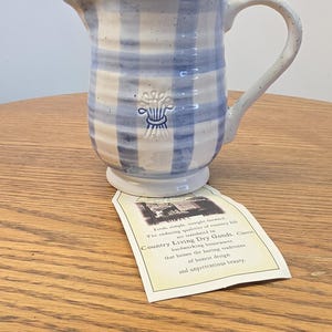 May include: A ceramic pitcher with a light blue and white checkered pattern, featuring a speckled finish and a small embossed wheat sheaf design. A paper tag is attached, printed with the text: Country Living Dry Goods. Classic hardworking housewares that honor the lasting traditions of honest design and unpretentious beauty.