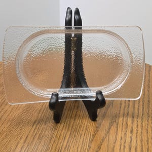 May include: A rectangular clear glass tray with rounded corners and a textured, pebbled surface finish. The tray features a central oval indentation and is displayed upright on a black plastic stand for clear visibility.