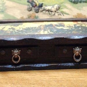 May include: A dark brown wooden box with a glass top, featuring a detailed landscape scene. The box has two small drawers with lion head ring pulls. The box is rectangular and sits on a wooden base.