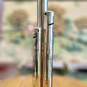 May include: A vintage brass and silver-toned cocktail jigger with three graduated measuring tubes. The jigger has etched markings for various measurements, including $1.00, $2.00, $3.00, $5.00, and $10.00. It sits on a round base.