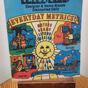 May include: A rectangular educational book titled Simplified Metrics for Everyday Use from Value City. The cover features a central sun illustration surrounded by four colorful panels depicting length, weight, volume, and temperature with cartoon characters.