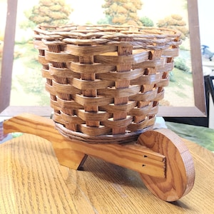 May include: A decorative, miniature wheelbarrow planter. The basket is woven from light brown material, and the wheelbarrow frame is made of light-colored wood. The wheel is a solid wooden circle. The planter is designed for small plants or decorative items.