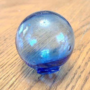 May include: A clear glass float with a blue base and accents. The round float has a small opening at the bottom and is sitting on a wooden surface. The float is a decorative item.
