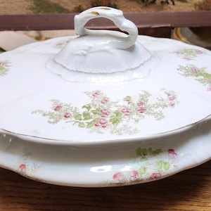 May include: White porcelain serving dish with lid. The dish features a floral pattern with pink and green accents. The lid has a decorative handle and the dish has gold trim. The dish is on a wooden surface.