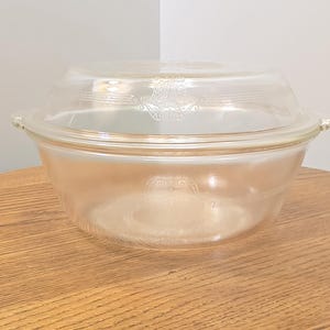 May include: A clear, light pink glass casserole dish with a matching lid. The bowl and lid feature a delicate, etched decorative pattern around the rim. The dish has two small side handles and a rounded base, presenting a translucent appearance.