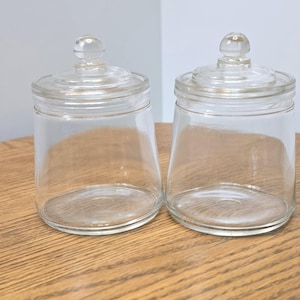 May include: Two clear glass canisters with matching removable lids. Each container features a cylindrical body with a slightly tapered base and a rounded knob handle on top of the lid. The glass is transparent and smooth throughout the entire structure.