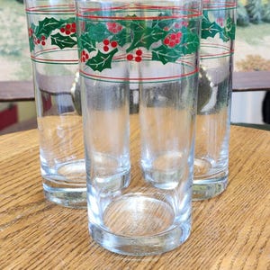 May include: Three clear glass tumblers featuring a decorative band near the top rim. The design consists of green holly leaves and red berries arranged in a horizontal pattern, accented by thin red and green lines encircling the glass.