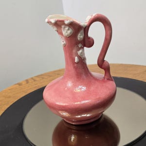 May include: A small pink ceramic pitcher with a glossy finish, featuring a curved handle and irregular white raised spots or drip-like textures concentrated around the rim and neck area, with a wide base and a narrow pouring spout.
