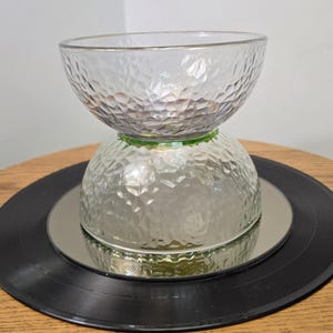 May include: A clear glass bowl with a textured, faceted exterior surface. The bowl is positioned upside down, resting on its rim, with a second identical clear glass bowl stacked on top of it, also oriented with its rim facing upward.