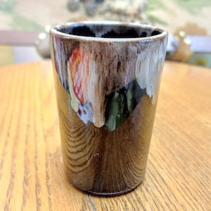 May include: A ceramic tumbler with a unique, textured glaze. The colors include shades of brown, white, red, and green, creating a marbled effect. The cup is cylindrical and appears to be handmade, suitable for beverages.