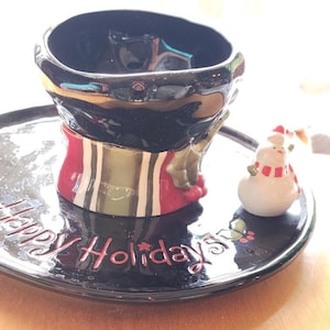 May include: A black ceramic mug and saucer set with a snowman figurine. The mug is black with a red, green, and gold design. The saucer has "Happy Holidays" in red script.