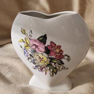 May include: White heart-shaped vase with a floral design. The flowers are in shades of pink, yellow, and red, with green leaves. The vase is approximately 6 inches tall.