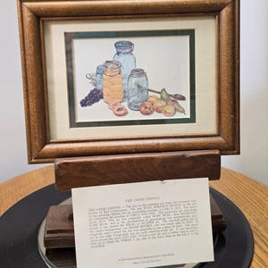 May include: A framed watercolor print titled THE GOOD THINGS depicting three glass canning jars, one filled with peaches, alongside grapes, pears, and a wooden spoon. The print is housed in a brown wooden frame with a cream mat and a dark green border.