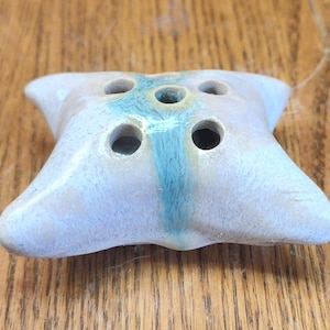 May include: A ceramic flower frog with a light blue and beige color scheme. The frog has a square shape with rounded edges and five holes for arranging flowers. A vertical stripe of turquoise blue runs down the center.