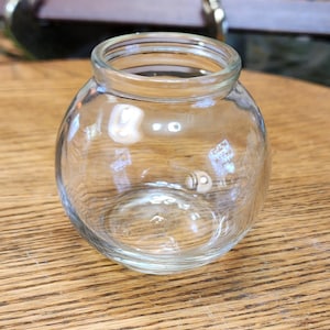 May include: A clear, round glass jar with a wide opening. The jar has a spherical body and sits on a wooden surface. The jar is empty and appears to be clean. The jar is suitable for storing small items or for decorative purposes.