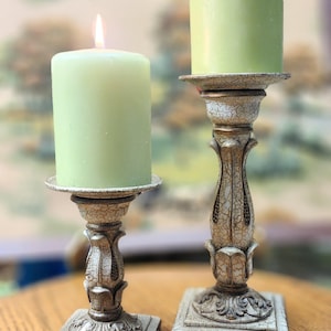 May include: Two decorative candle holders with green pillar candles. The candle holders have a distressed, off-white finish with ornate detailing. One candle is lit. The candle holders are on a wooden surface.
