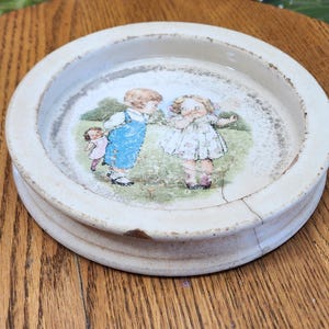 May include: A round, off-white ceramic dish with a raised rim. The center features a colorful illustration of three children in a grassy field. The dish has visible cracks and signs of age, adding to its vintage charm.
