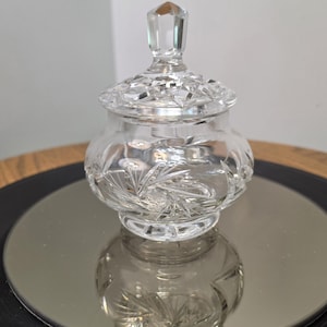 May include: A clear, colorless glass candy dish with a matching lid. The vessel features intricate, geometric cut-glass patterns, including starburst motifs on the body and a faceted, diamond-shaped finial handle atop the circular lid.