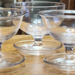 May include: Three clear glass dessert dishes with a stemmed base and a round, flat foot. The bowls are round with a slight flare at the top. The dishes are sitting on a wooden surface.