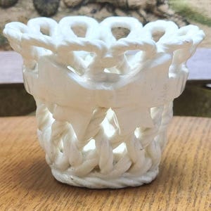 May include: A white, decorative basket-style container with a woven rope design. The container has a cylindrical shape with a flared top and a banner-like detail on the side. The basket is made of a solid, opaque material.