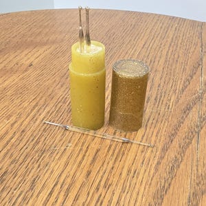 May include: A cylindrical yellow container with a speckled finish holds two thin metal tools with small spherical tips. Beside it sits a separate, matching cylindrical cap with a gold glitter finish. A third thin metal tool lies horizontally.