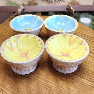 May include: Four ceramic flower-shaped bowls. Two bowls are light blue, and two are yellow. The bowls have a textured, lattice-like exterior and sit on a pedestal base. The bowls are arranged on a wooden surface.