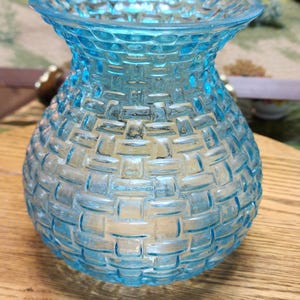 May include: A light blue glass vase with a woven basket design. The vase has a rounded body and a flared rim. The glass is transparent, allowing light to pass through. The vase is sitting on a wooden surface.