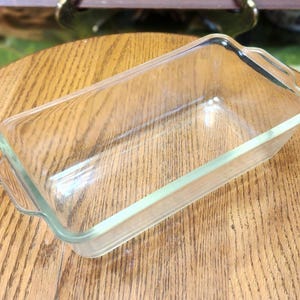 May include: A clear, rectangular glass loaf pan with rounded corners and small handles on each end. The pan is empty and sits on a wooden surface. This baking dish is ideal for making bread, cakes, and other baked goods.