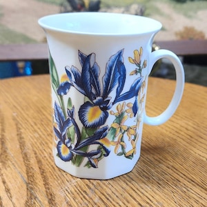 May include: White ceramic mug with a floral design featuring blue irises and yellow flowers. The mug has a hexagonal shape and a curved handle. The floral pattern is set against a cream-colored background.