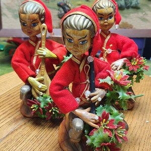 May include: Three vintage figurines of Asian-inspired figures in red hooded outfits. Each figure has a painted face, gold-colored hair, and is holding a musical instrument or decorative item. The figurines are decorated with red and green floral accents.