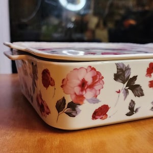 May include: A rectangular ceramic baking dish with a lid. The dish is cream-colored and decorated with red and pink floral patterns and dark green leaves. It has two small handles on the sides.