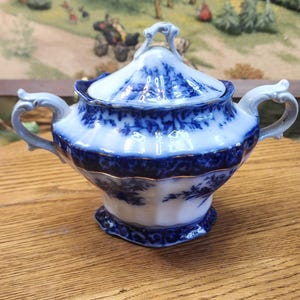 May include: A blue and white porcelain sugar bowl with a lid. The bowl has two handles and a decorative floral pattern. The rim and base are trimmed with gold. The sugar bowl is sitting on a wooden surface.