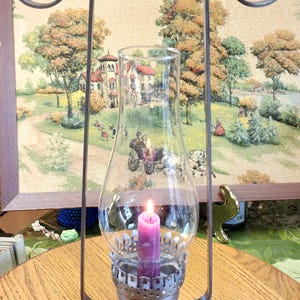 May include: A vintage metal and glass oil lamp with a lit purple candle inside. The lamp has a decorative metal frame and base. The background features a framed tapestry with a pastoral scene.