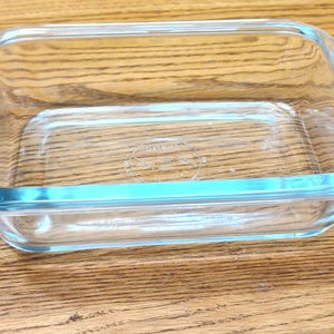 May include: A clear, rectangular Pyrex baking dish with a light blue rim. The dish is empty and sits on a wooden surface. The Pyrex logo is visible on the bottom of the dish.