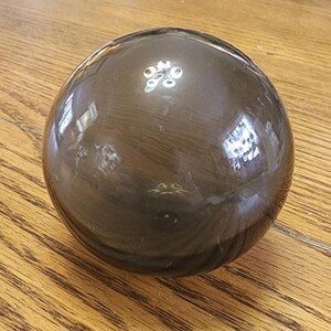 May include: A dark, glossy, spherical object with a reflective surface. The object appears to be made of a dark, possibly smoky, glass or crystal. The sphere is sitting on a wooden surface.